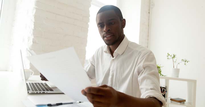 Hombre mirando seriamente dos hojas de papel encima de la computadora portátil y la pluma en el escritorio blanco