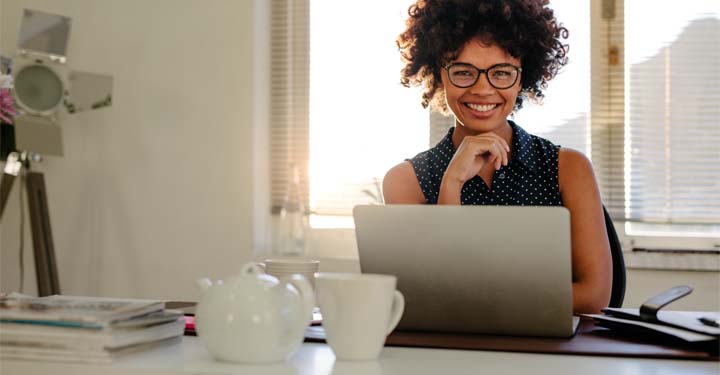 Mujer en la oficina en casa hace una pausa del trabajo para sonreír con la mano debajo de la barbilla con tetera frente a su computadora portátil
